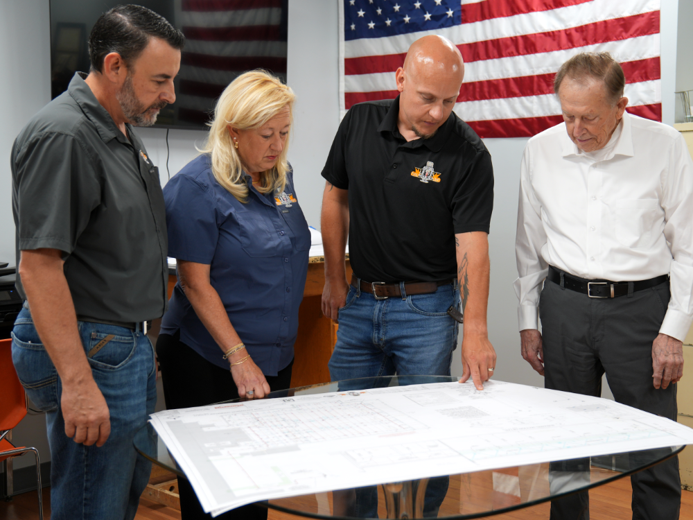 Employees and owner, Walter Lewis, gathered around a table, pointing at a blueprint, while discussing a project.